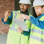 Two construction managers in safety vests and hard hats reviewing project plans on a tablet, demonstrating improved digital communication between a contractor and subcontractor.